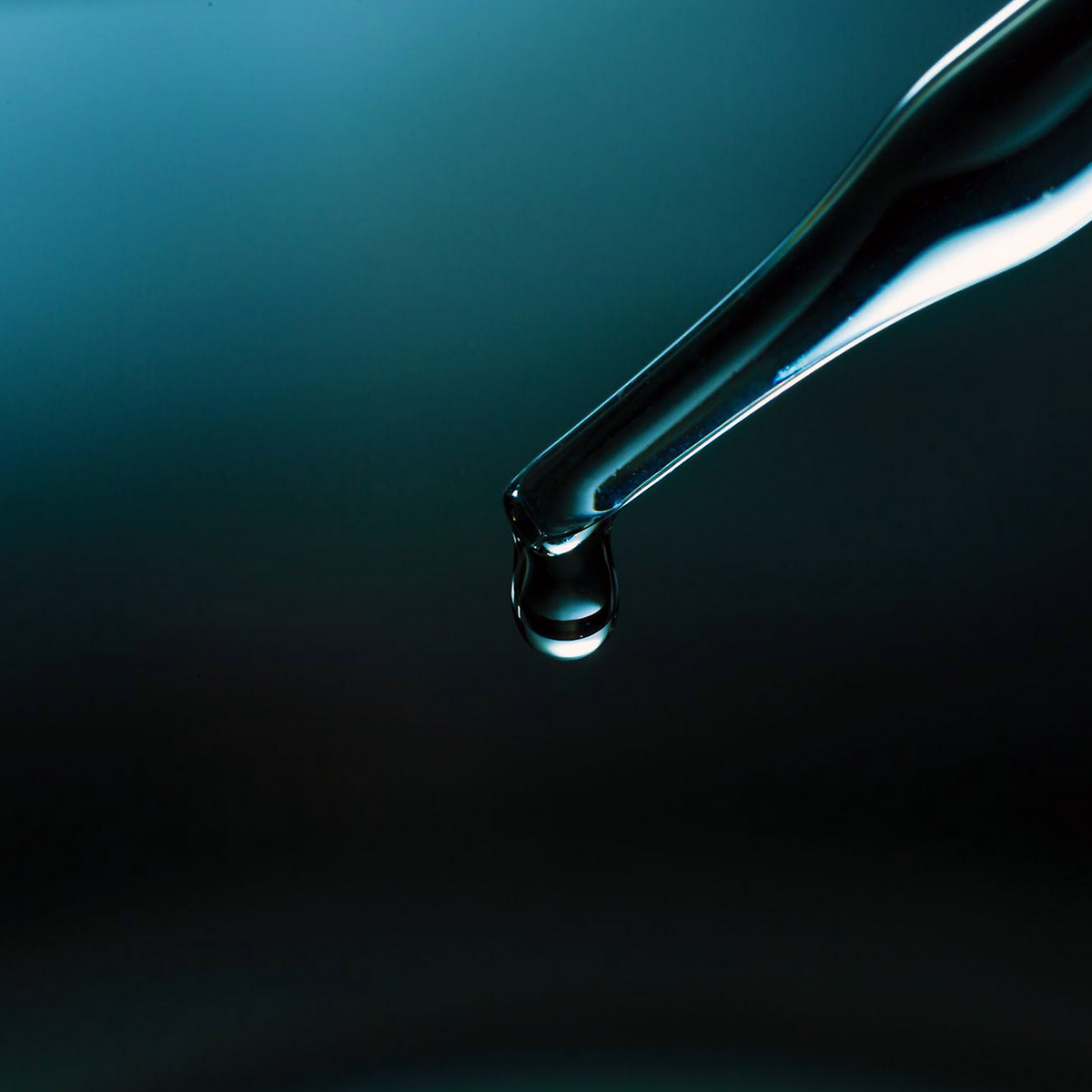 Egyptian Blue Lotus Hydrosol, clear droplet hanging from a glass pipette entering from the upper right against a blue-black background
