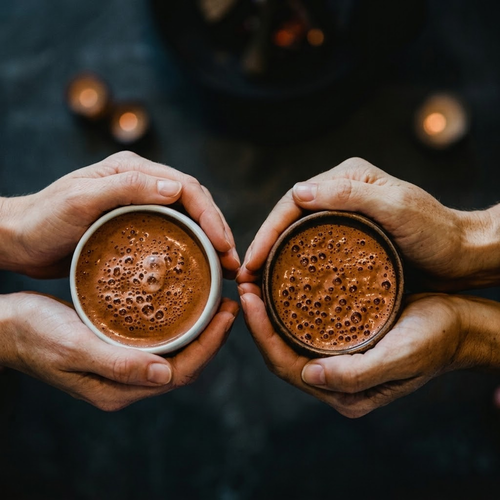 Two people holding cups of frothy ceremonial cacao against a dark background with warm candlelight